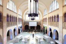 Großer, heller Veranstaltungssaal mit hohen Decken, Stehtischen und Leinwand.