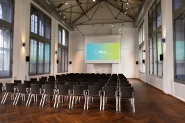 Leerer Konferenzraum mit Stuhlreihen und großer Leinwand vor hohen Fenstern