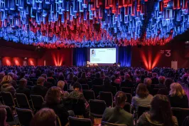 Großer Konferenzsaal mit vielen Zuschauern, Bühne vorne, bunte Deckenbeleuchtung.