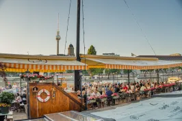 Sonnige Terrasse von Spree Fumée, Markisen spenden Schatten, zahlreiche Gäste genießen das Ambiente bei strahlendem Wetter.