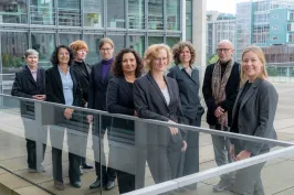 Neun Personen in Businesskleidung stehen draußen vor modernen Bürogebäuden.