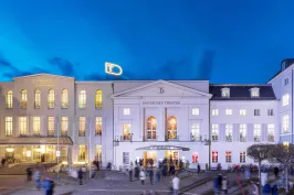 Beleuchtetes Theatergebäude abends mit Menschen auf dem Platz davor.