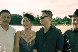 Vier Erwachsene stehen lächelnd am Wasser, sommerliche Kleidung, Bäume im Hintergrund.