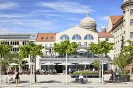 Belebter Platz mit Bäumen und Café vor historischem Gebäude mit Kuppel.