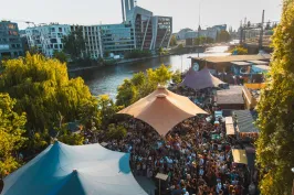 Belebtes Open-Air-Gelände vom Holzmarkt mit Zeltdächern und Menschenmenge am Spreeufer in der Abendsonne, im Hintergrund Stadtgebäude.