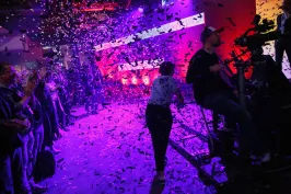 Bühne mit lila Licht, Konfetti fällt, Publikum applaudiert, Kameramann filmt.