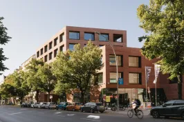 Modernes Backsteingebäude an einer Straße mit Bäumen, Autos und einem Radfahrer.