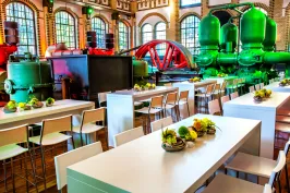 Modern dining tables with floral centerpieces in a colorful industrial hall with large machinery.