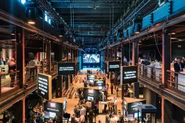 Große Messehalle mit Ständen, Besuchern und einer beleuchteten Bühne im Hintergrund.