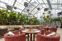 Restaurant mit runden Tischen, roten Sesseln und viel Grün unter Glasdach.