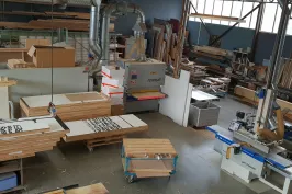 Holzwerkstatt mit Maschinen, Holzplatten und Werkzeugen, Tageslicht durch Fenster.