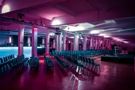 Reihe leerer Stühle in beleuchtetem Veranstaltungssaal mit Bühne und buntem Licht.