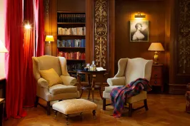 Gemütliche Bibliothek mit zwei Sesseln, rotem Vorhang, Bücherregal und warmem Licht.