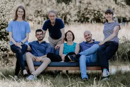 Sechs lächelnde Menschen sitzen und stehen auf einer Bank im Freien.