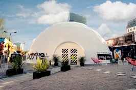 Weißes Kuppelzelt mit Pflanzen und bunten Stühlen auf einem Platz, blauem Himmel im Hintergrund.