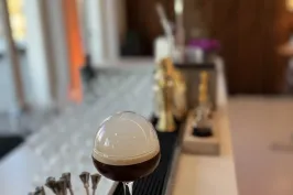 Cocktailglas mit Schaumkrone auf Bartheke, im Hintergrund Gläser und Barutensilien.