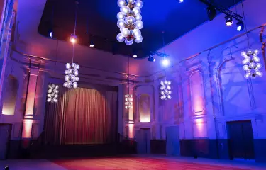 Der große Festsaal des Peter Edel mit Effektbeleuchtung, man sieht den Blick auf die Bühne, einen Bühnenvorhang sowie die neobarocken Zierelemente an den Wänden und die Deckenlampen, die im Stil des Palastes der Republik gehalten sind