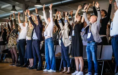 Viele Menschen stehen von ihren Stühlen auf und strecken ihre Arme in die Höhe.