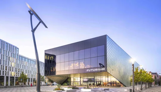 Modernes, würfelförmiges Gebäude mit Glasfassade auf einem gepflasterten Platz bei Tageslicht.