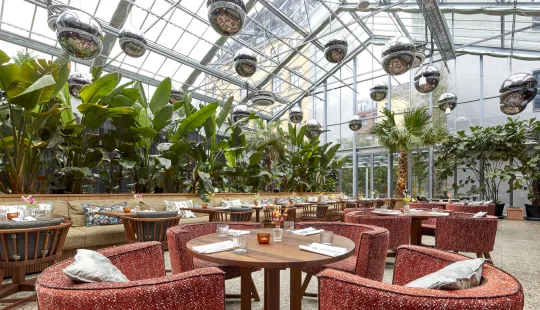 Helles Restaurant mit Pflanzen, Glasdach und gemütlichen Sesseln.