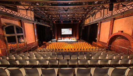 Leerer Veranstaltungsraum mit Bühne und vielen Reihen orangefarbener Stühle.