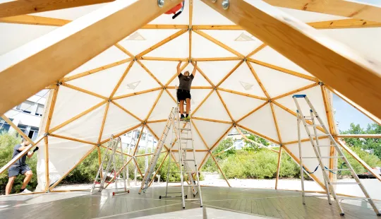 Person steht auf einer Leiter unter einem halb fertigen Holzkuppel-Zelt im Freien.