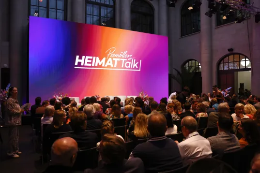 Zuschauer sitzen vor einer bunten Bühne mit „HEIMATtalk” Schriftzug.