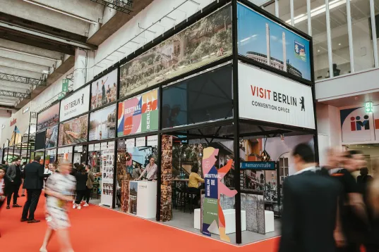 Visitors walk through the exhibition hall past the colorful visitBerlin booth featuring images of the city.