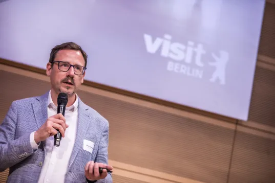 Mann mit Mikrofon präsentiert vor „visit Berlin“-Logo.