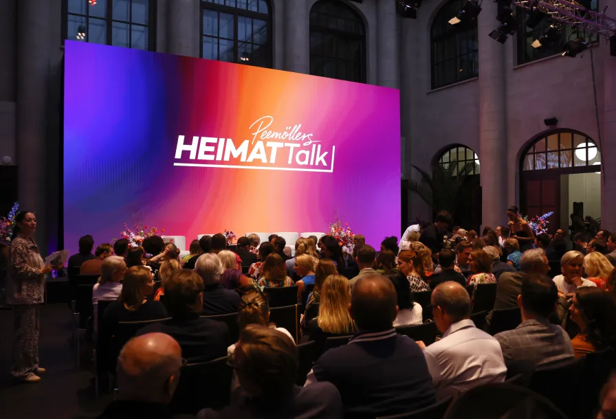 Audience seated in front of a colorful stage with "Heimat Talk" displayed on a large screen.