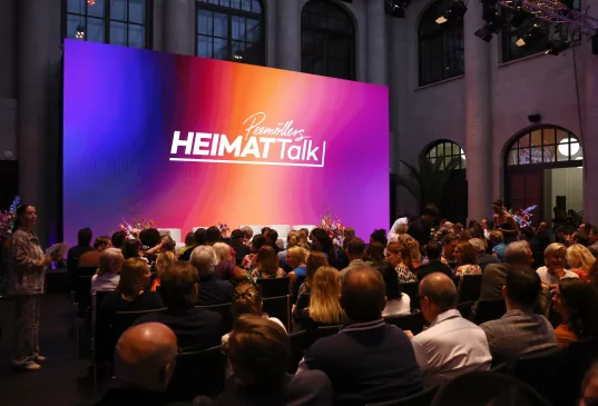 Zuschauer sitzen vor einer bunten Bühne mit „HEIMATtalk” Schriftzug.