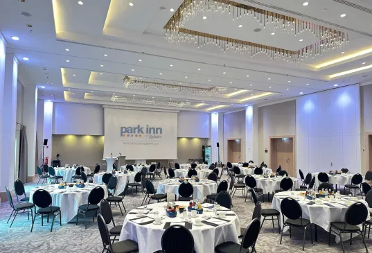 Banquet hall with round tables set for an event, white tablecloths, stage at front.