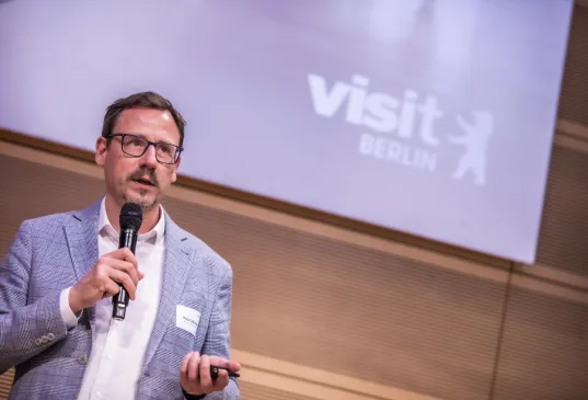 Man holding a microphone speaking, "visit Berlin" projected on screen behind him.