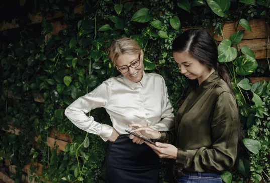 Two women in business attire stand in front of an ivy-covered wall and look at an Tablet with smiles on their faces.