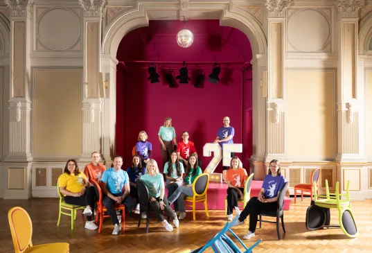 Group of people in colorful shirts pose on chairs in a grand hall with a large "25" sign.