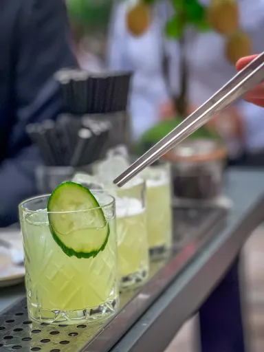 Cocktail with ice and cucumber slice garnish on a bar counter, with a metal straw above.