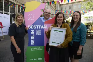 Vier Personen stehen nebeneinander vor einer bunten BESTIVAL-Bärenskulptur. Eine Frau hält stolz ein eingerahmtes Zertifikat (ISO 20121) in der Hand. Im Hintergrund sind bunte Wimpel, andere Besucher*innen und ein sonniger Innenhof zu sehen.