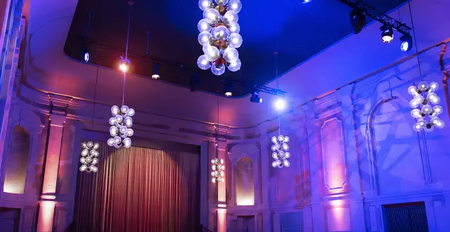 Der große Festsaal des Peter Edel mit Effektbeleuchtung, man sieht den Blick auf die Bühne, einen Bühnenvorhang sowie die neobarocken Zierelemente an den Wänden und die Deckenlampen, die im Stil des Palastes der Republik gehalten sind