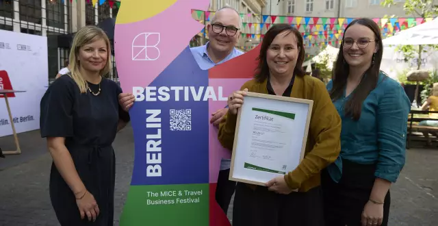 Vier Personen stehen nebeneinander vor einer bunten BESTIVAL-Bärenskulptur. Eine Frau hält stolz ein eingerahmtes Zertifikat (ISO 20121) in der Hand. Im Hintergrund sind bunte Wimpel, andere Besucher*innen und ein sonniger Innenhof zu sehen.