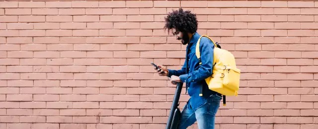 Young man with backpack and E-Scooter standing in front of brick wall looking at cell phone