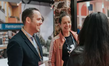 Zwei Personen schauen lächeln eine dritte Person angucken, die mit dem Rücken zur Kamera steht.