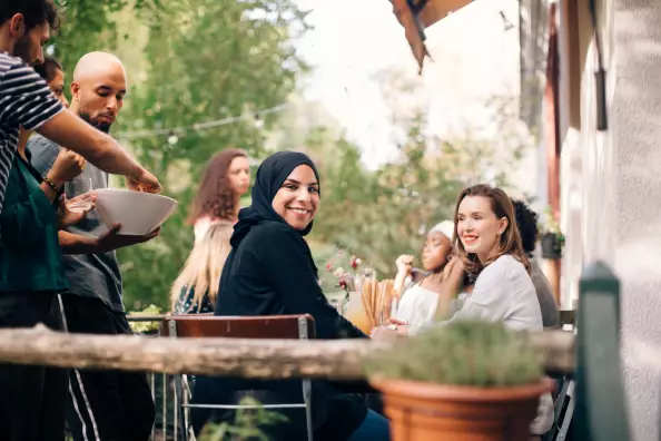 Porträt einer jungen Frau mit Hijab, die während einer Party mit Freunden auf dem Balkon sitzt und lächelnd in die Kamera schaut