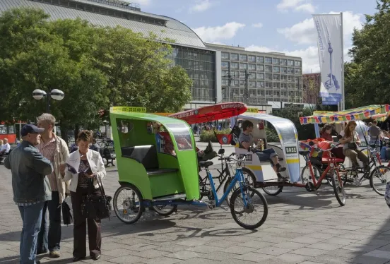 Mehrere bunte Fahrradrikschas stehen auf einem Platz, im Hintergrund Bäume und Gebäude.
