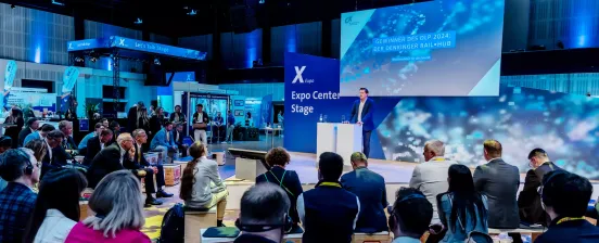 A speaker on stage at X Expo, with an interested audience seated in front of him.