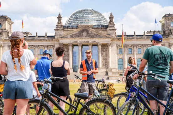 A group of cyclists stands in front of the Reichstag with a group leader.