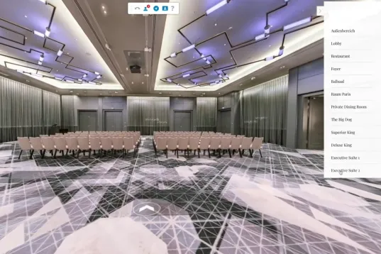 Modern conference room with rows of chairs, patterned carpet, and video call windows in the corner.