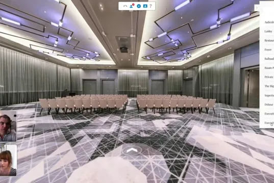 Modern conference room with rows of chairs, patterned carpet, and video call windows in the corner.