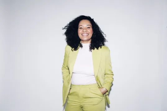 a picture of the founder and Managing Director of The Impact Company, Yolanda Rother, in a bright green suit, smiling, she's standing in front of a white wall