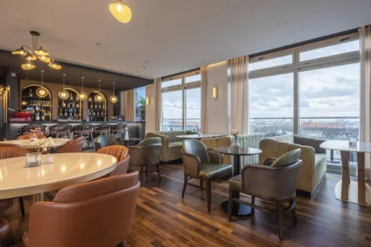 Modern lounge with large windows, cushioned chairs, and a bar area in the background.