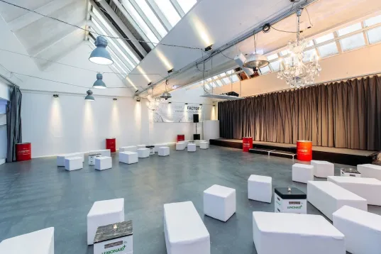 Bright event room with white seating cubes and stage, large windows through the shed roof and curtains.
